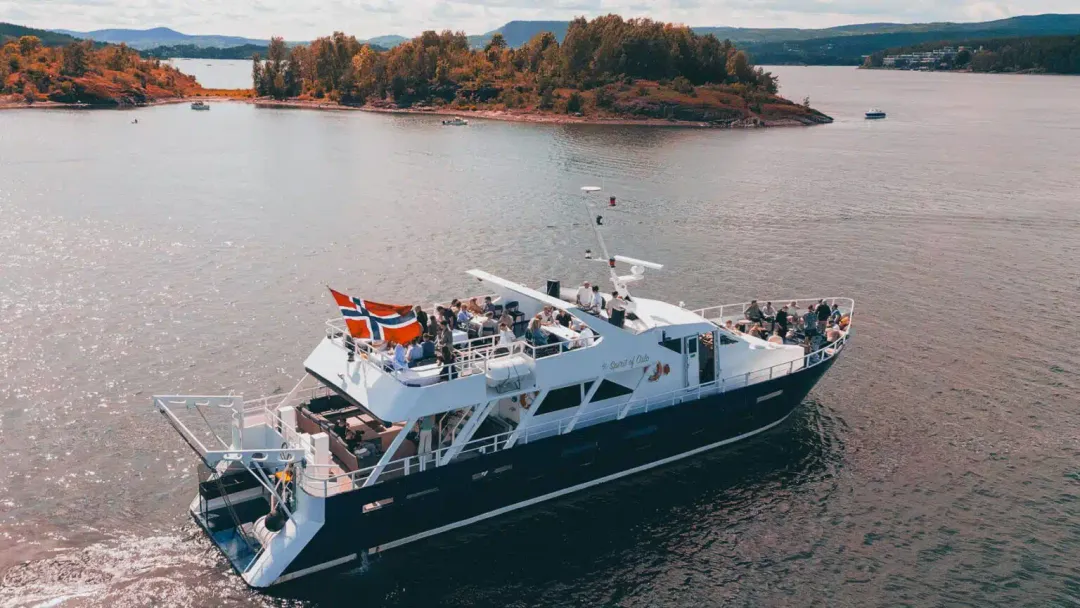 Båten My Spirit of Oslo på fjorden med gjester på soldekket og norsk flagg akterut